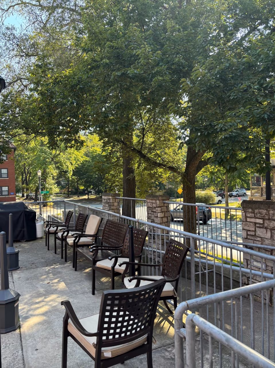 Outdoor patio area with several cushioned metal chairs arranged along a concrete walkway next to a metal railing. Large leafy trees provide shade, and a street with parked cars and residential buildings is visible in the background.
