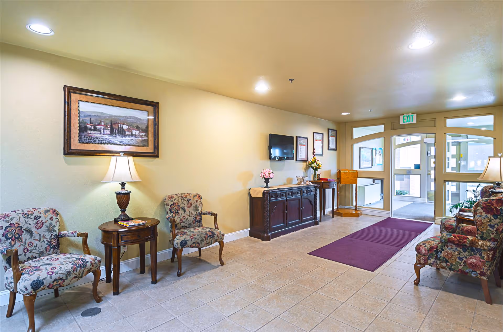 Well-lit lobby with floral upholstered chairs, side tables, a console cabinet, and glass entry doors.