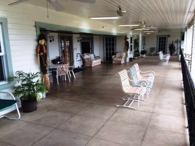 A covered outdoor patio area with several white metal rocking chairs and wicker furniture arranged along the wall. The patio has a tiled floor, ceiling fans, and decorative scarecrow figures near the doors. There are potted plants and a railing along the open side of the patio.