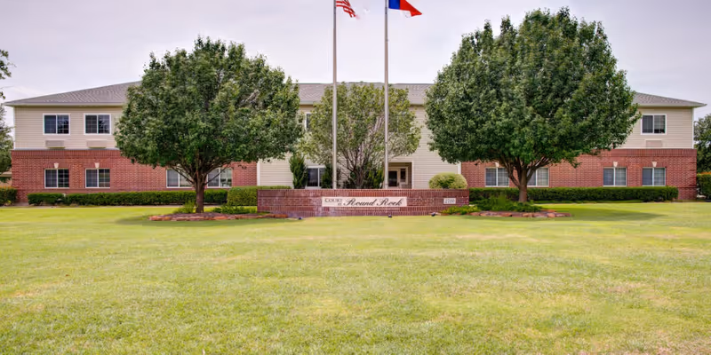 Front exterior view of The Court at Round Rock building with two large trees, a well-maintained lawn, and two flagpoles displaying the American and Texas flags.