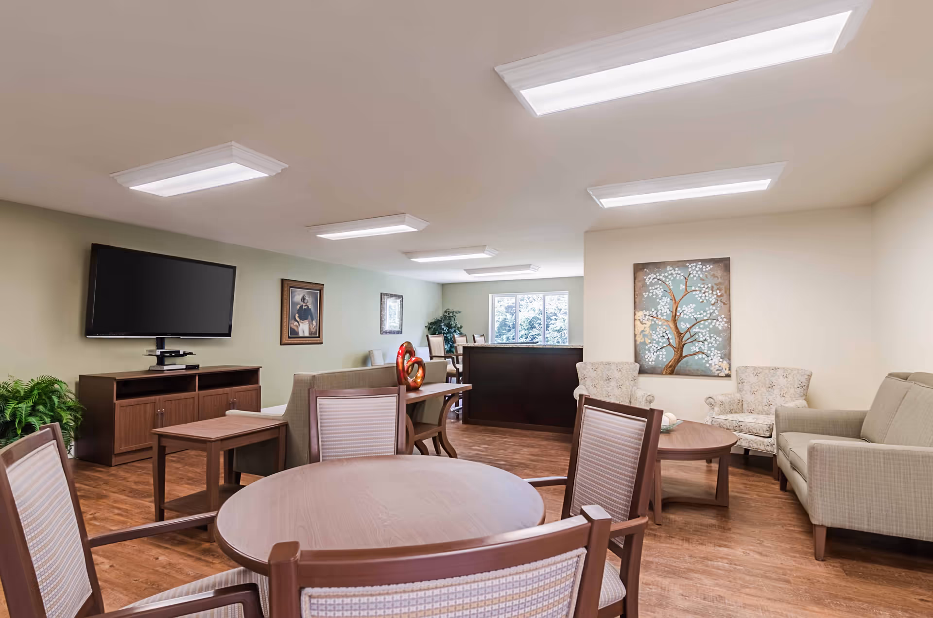 A well-lit common area in a senior living facility featuring a round wooden table with four chairs in the foreground, a seating area with a sofa, two armchairs, and a coffee table to the right, a flat-screen TV mounted on a wooden cabinet against a green wall on the left, and a dining area with chairs and a window in the background. The room has wood flooring, ceiling lights, framed artwork on the walls, and a decorative plant near the TV.