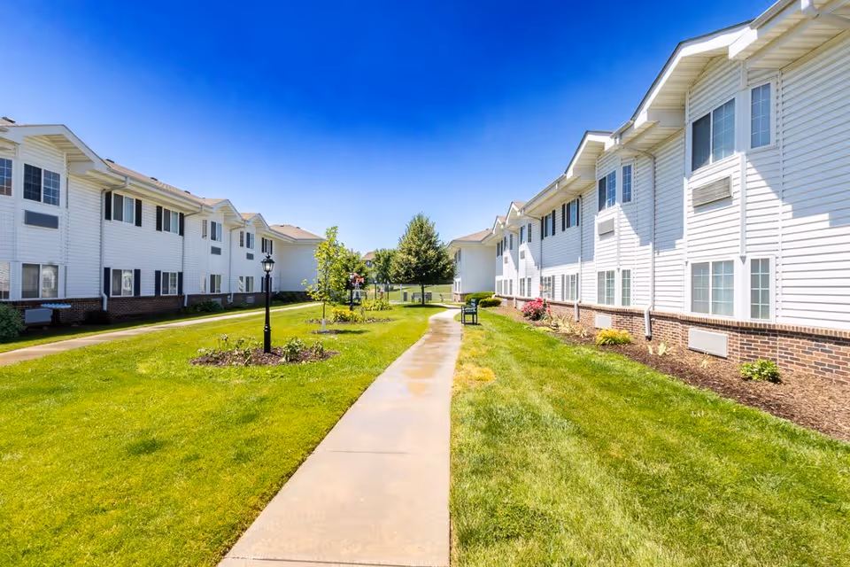 A paved walkway runs between two white two-story residential buildings with brick foundations. The area is landscaped with green grass, small trees, flower beds, and benches under a clear blue sky.