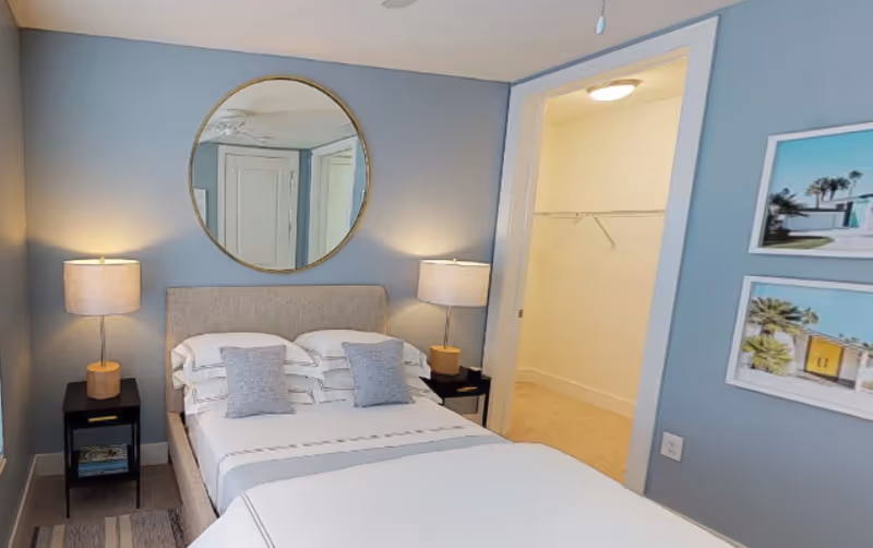 A neatly made bed with a beige upholstered headboard and two gray pillows in a bedroom with light blue walls. Two black nightstands with wooden lamps are on either side of the bed. A large round mirror hangs above the bed. To the right, there is an open doorway leading to a walk-in closet with white walls and a beige carpet. Two framed pictures of outdoor scenes hang on the wall next to the closet.