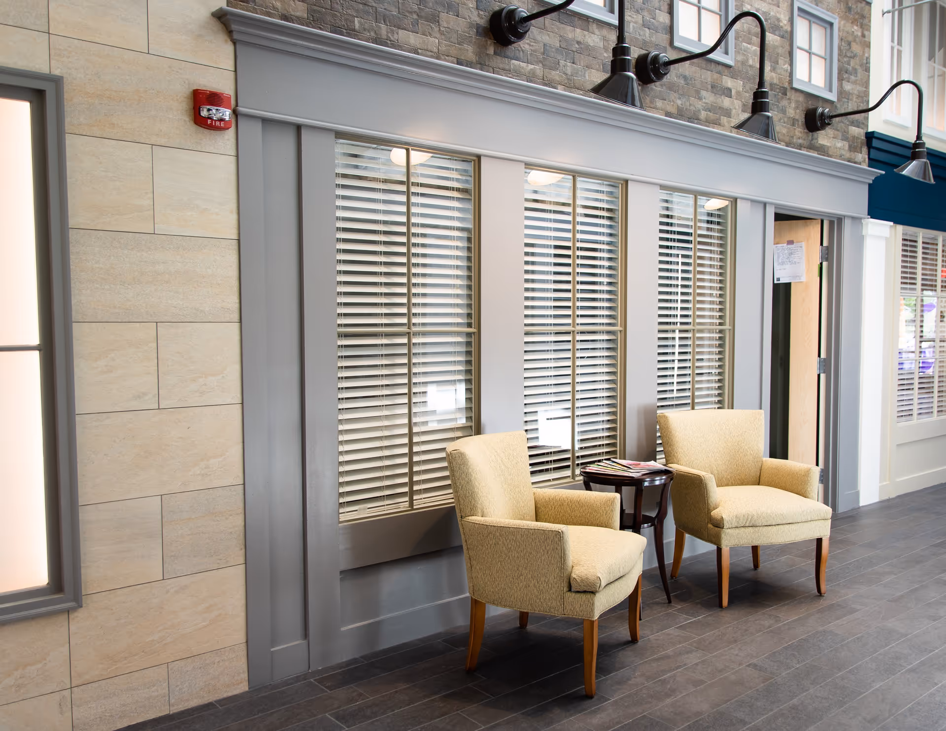 Two beige armchairs and a small round table placed in a hallway-style interior seating area against windowed wall panels.