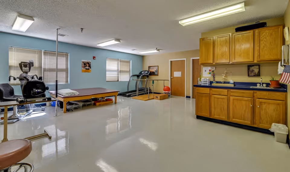 A spacious therapy or rehabilitation room with exercise equipment including a stationary bike, treadmill, and therapy table. The room has light blue and beige walls, two windows with blinds, wooden cabinets with a countertop, and a clean, shiny floor.