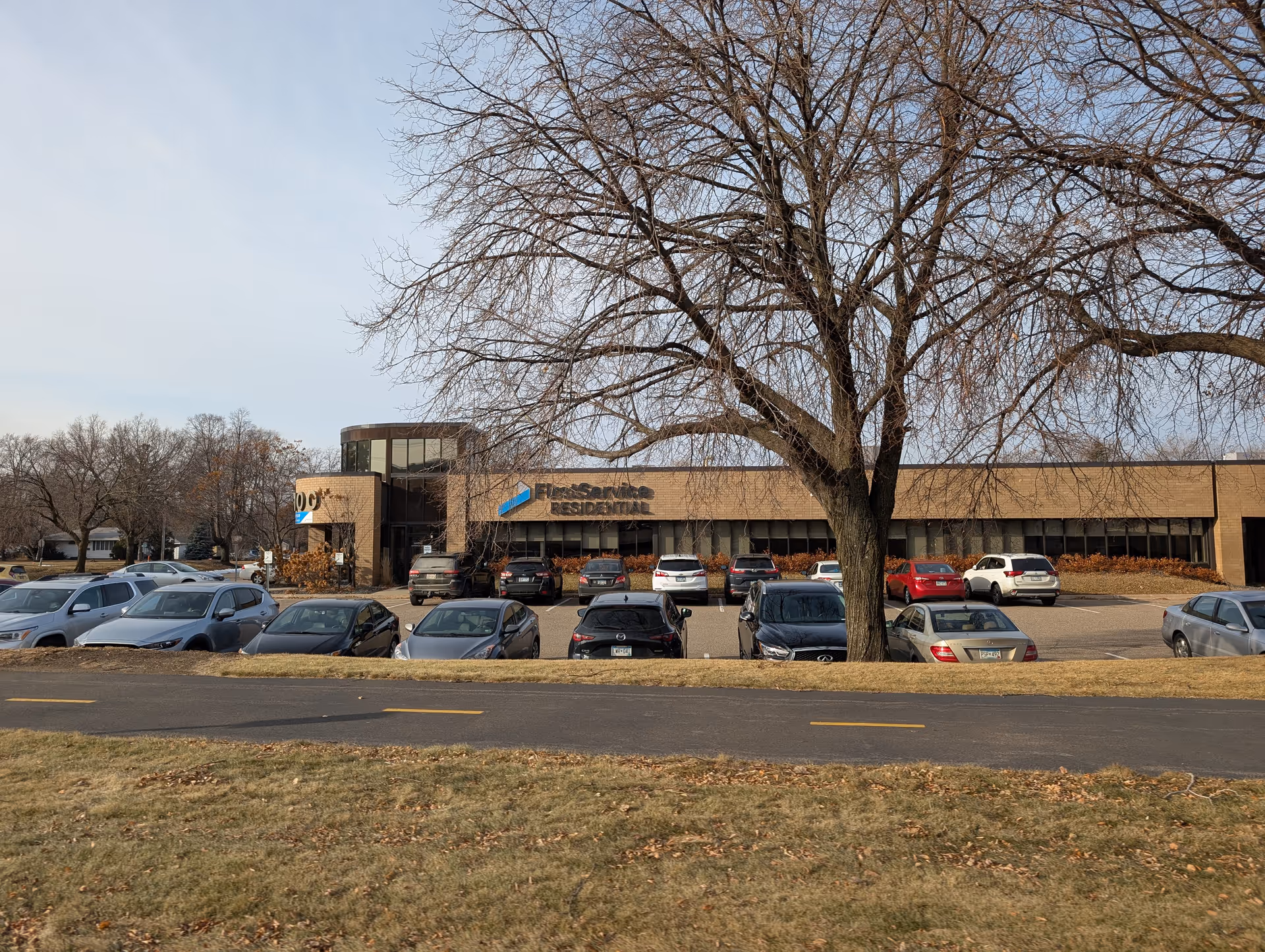 Exterior view of a single-story commercial building with a parking lot in front. Several cars are parked, and there is a large leafless tree in the foreground. The building has a sign that reads 'FlexService RESIDENTIAL'. The sky is clear with some light clouds.