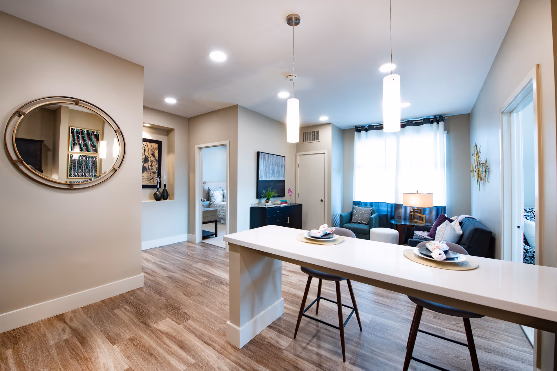 Interior view of a senior living facility apartment showing a modern open living area with a kitchen island and two stools in the foreground, a living room with a sofa, armchair, and lamp near a window with curtains, and two bedrooms visible through open doorways. The space features wood flooring, neutral walls, and contemporary lighting fixtures.