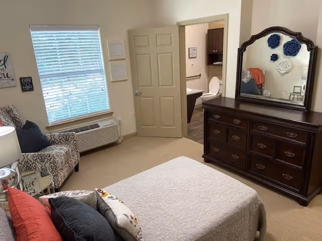 A bedroom with a bed covered in a light-colored quilt and multiple pillows. There is a patterned armchair near a window with blinds, and a wooden dresser with a mirror on the right side. The door to an adjoining bathroom is open, showing a toilet and part of a sink.