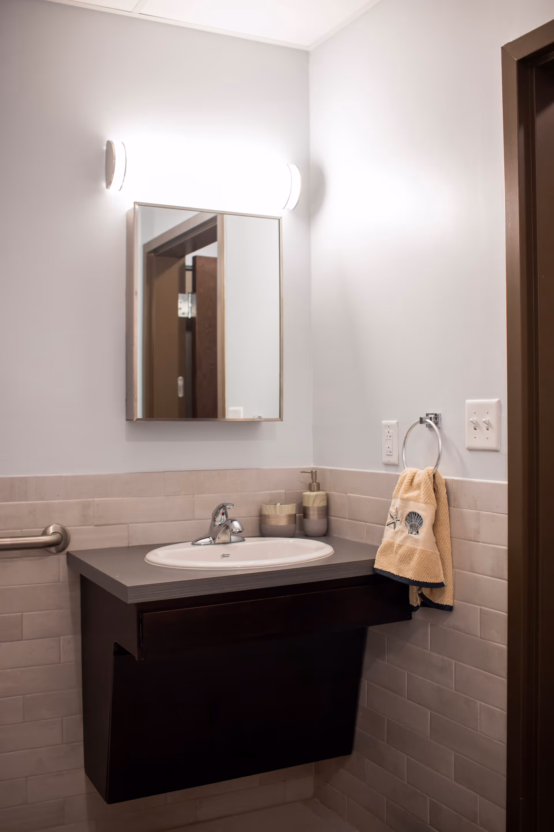 A clean bathroom sink area with a dark wood vanity, a white oval sink, a chrome faucet, a wall-mounted mirror with a bright light above it, beige tiled walls, a towel ring holding a beige towel with seashell designs, and light switches on the wall.