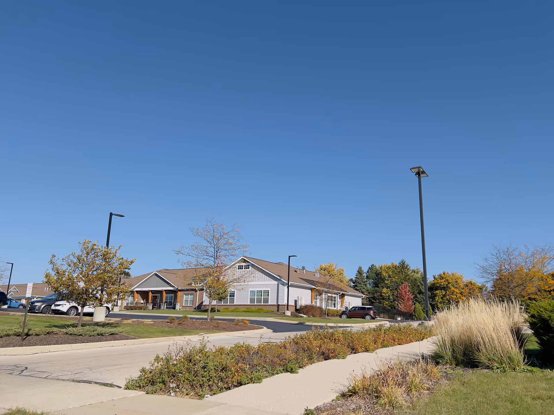 Exterior view of a single-story senior living facility building with a parking lot, trees, shrubs, and a clear blue sky.