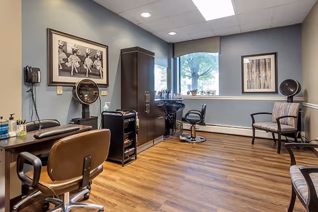 A small salon room with styling chairs, hooded hair dryers, a storage cabinet, and waiting chairs by a window.