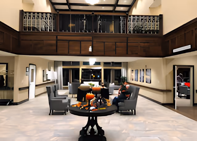 A spacious and well-lit common area in a senior living facility with high ceilings and a mezzanine balcony. The room features comfortable armchairs arranged around a central round table decorated with autumn-themed items including a pumpkin and leaves. There are a few people seated and conversing, and the walls are adorned with framed pictures. The area has a warm and inviting atmosphere.