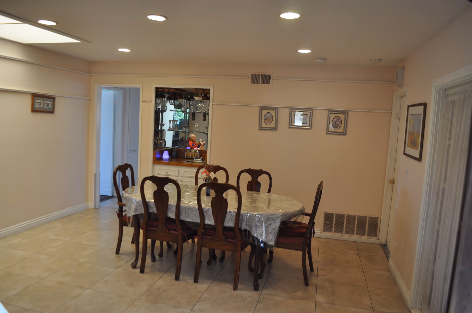A dining room with a rectangular wooden table covered with a lace tablecloth and six matching wooden chairs with red cushions. The room has beige walls with three framed pictures hanging on one wall and a small built-in cabinet with glassware and a mirror. The floor is tiled, and there is a door and a window with sheer curtains on the right side.