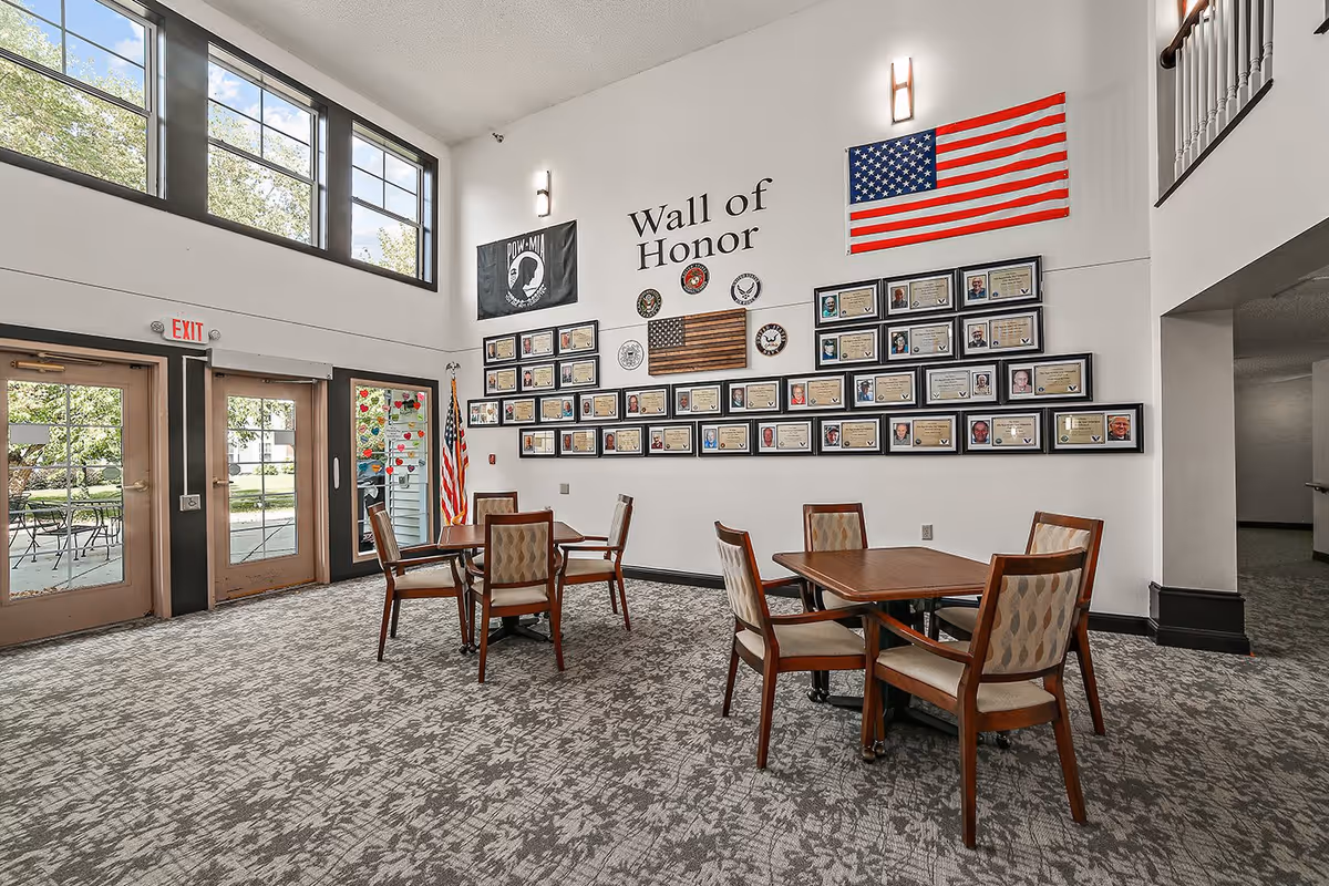 Interior view of a common area in The Rivers Retirement Community featuring a Wall of Honor with framed photos and certificates, an American flag, and a POW-MIA flag. The room has high ceilings, large windows, carpeted floor, and several wooden tables with chairs. Double glass doors lead outside to a patio area with outdoor seating.