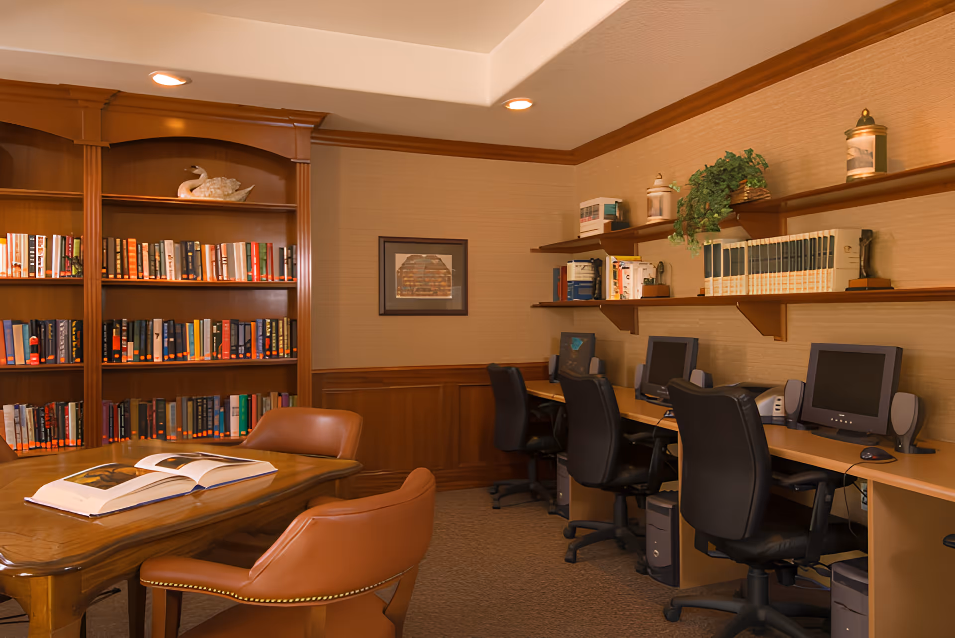 Cozy communal library and computer room with bookshelves, a wooden table with open books, and a row of desktop computers and chairs.