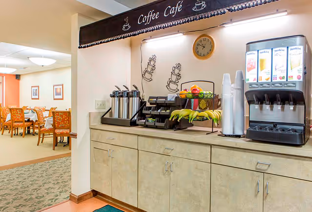 A coffee station in a senior living facility with coffee dispensers, a juice dispenser, cups, bananas, and other fruit on a countertop. In the background, there is a dining area with tables and chairs. A sign above the station reads 'Coffee Cafe' with coffee cup illustrations and a wall clock showing the time.