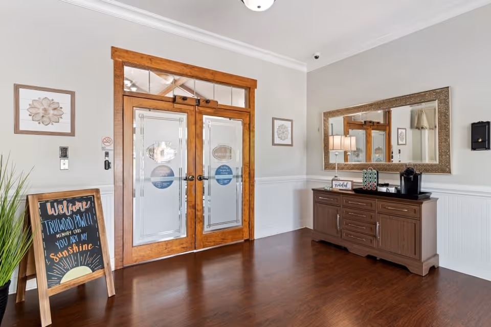 Interior view of a facility entrance area with double wooden doors featuring frosted glass panels. To the right is a wooden sideboard with a coffee maker, lamp, and decorative items, and above it hangs a large ornate mirror. To the left, there is a chalkboard sign with a welcoming message and a potted plant beside it. The floor is wooden, and the walls are painted light gray with white wainscoting.