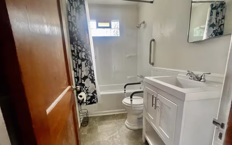 Small bathroom with a tub/shower and floral curtain, a toilet fitted with safety arms, white vanity sink, and a grab bar.