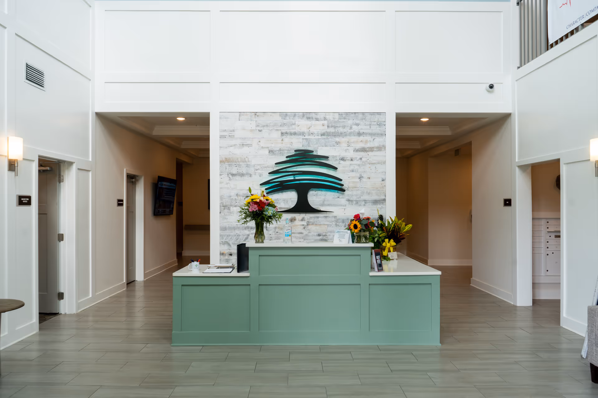 Reception area of The Canopy at Hickory Creek featuring a green front desk with flower arrangements on top, a large stylized tree logo on a whitewashed wooden wall behind the desk, and hallways on either side leading to other parts of the facility.