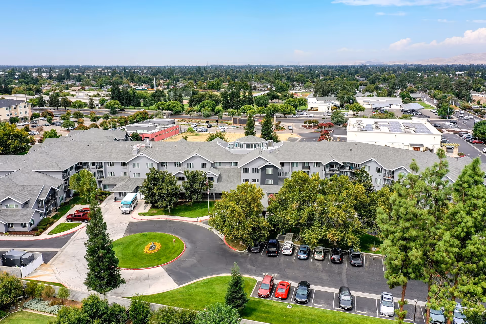 Aerial view of Solstice Senior Living at Clovis, showing a large multi-story residential building surrounded by trees, green lawns, and a parking lot with several cars. The building has a gray roof and light-colored exterior walls. The surrounding area includes other buildings, roads, and greenery under a clear sky.