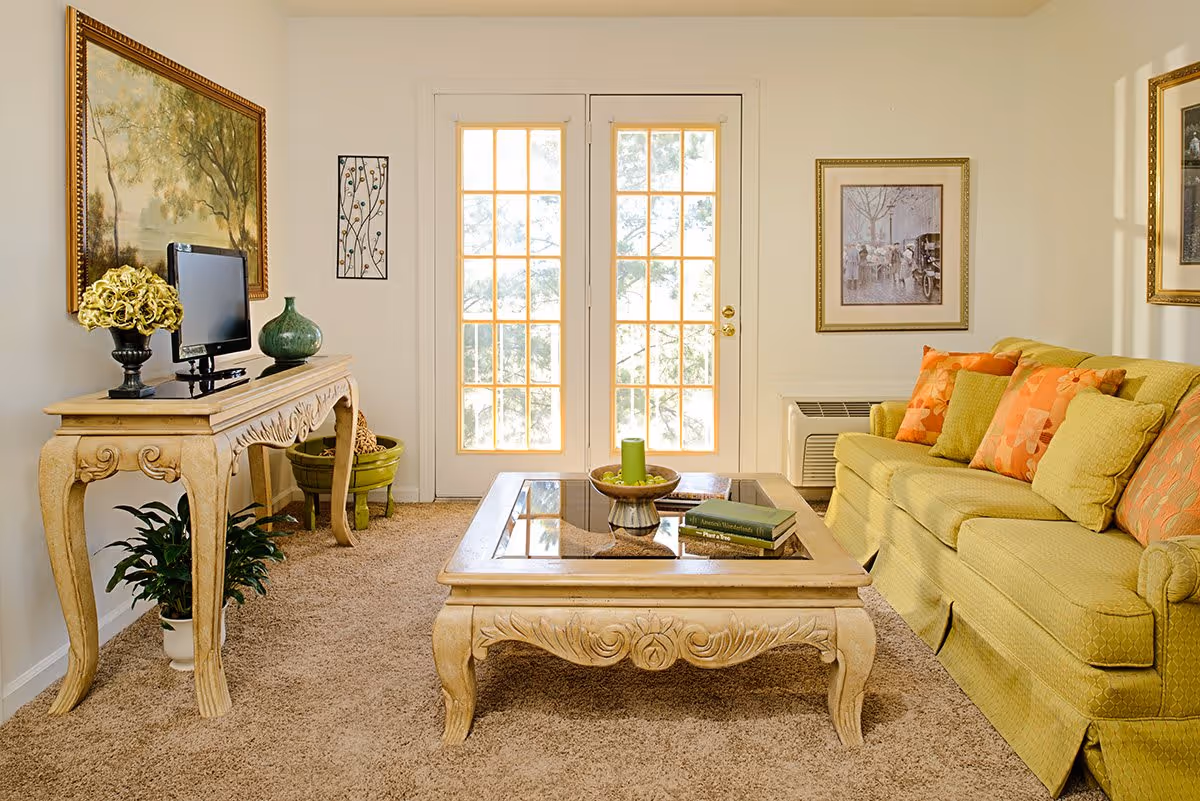 A cozy living room with a green sofa adorned with orange and green patterned cushions. In front of the sofa is a wooden coffee table with a glass top, holding a green candle and two books. To the left, there is a decorative wooden console table with a small flat-screen TV, a green vase, and a flower arrangement. The room has beige carpet flooring and light-colored walls with framed artwork. French doors with glass panes allow natural light to enter the room.