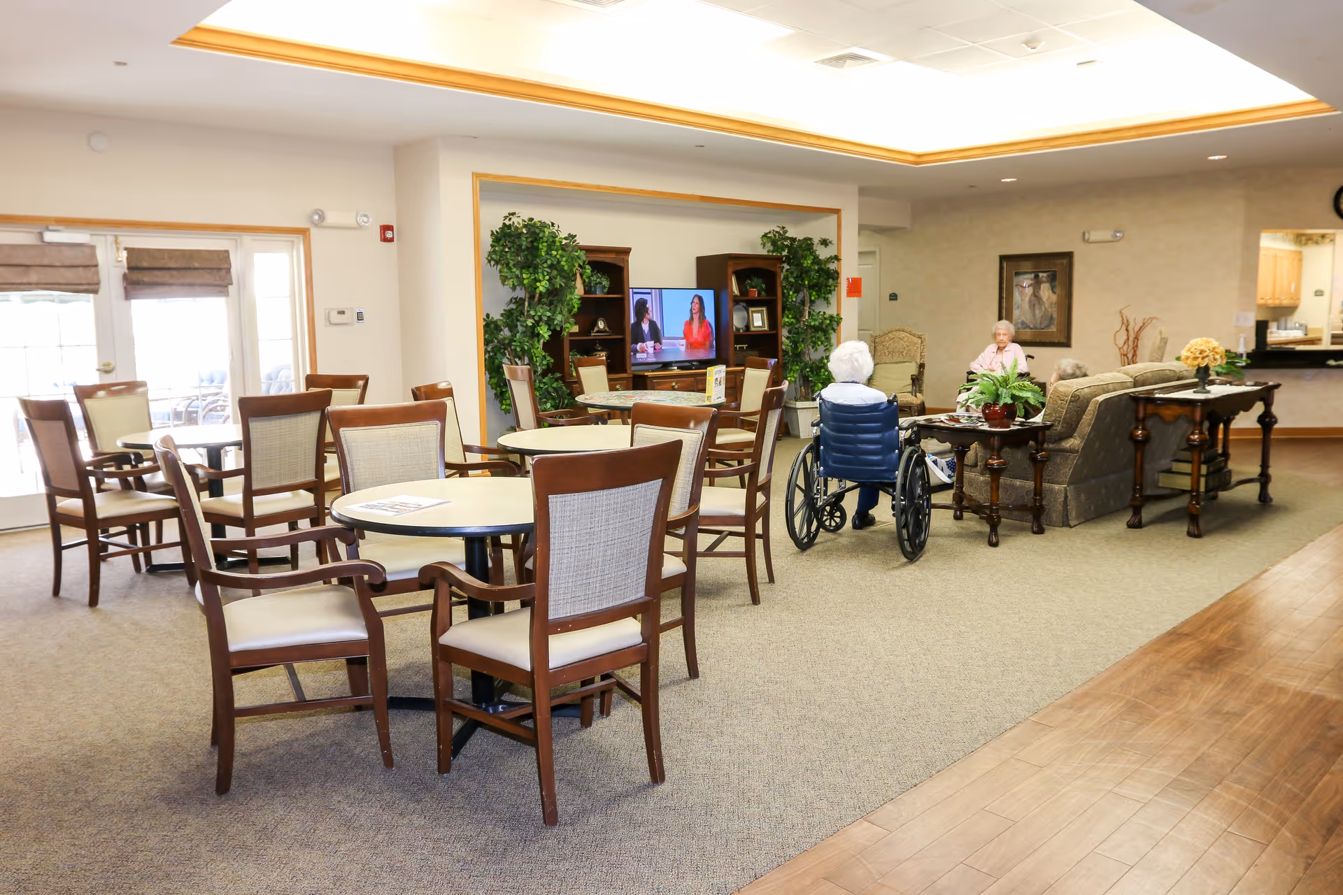 A spacious senior living common area with several round tables and chairs arranged for seating. Two elderly women are seated near a couch, one in a wheelchair, watching a television mounted on a wooden entertainment center with plants on either side. The room has carpeted flooring with a wooden section on the right and large windows letting in natural light.