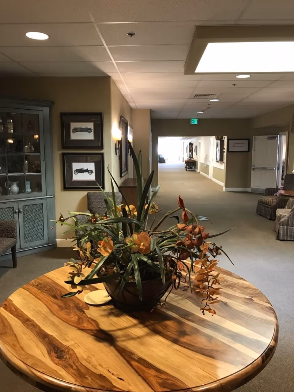 A hallway in an assisted living facility with a round wooden table in the foreground holding a large floral arrangement with green leaves and orange flowers. The hallway has beige walls, carpeted floors, framed pictures on the walls, and several chairs along the sides. There is a cabinet with glass doors on the left side and an exit sign visible at the far end of the hallway.