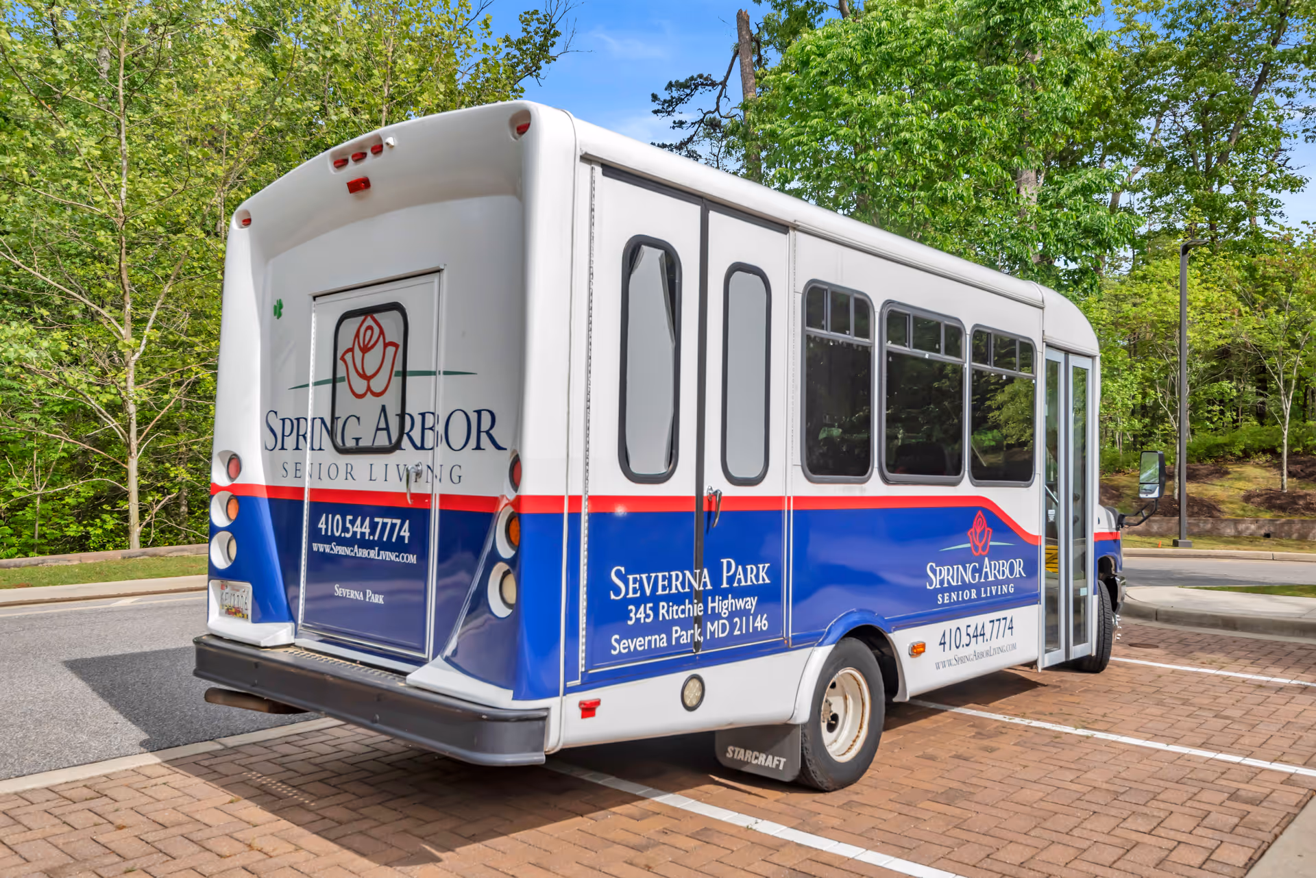 A white and blue shuttle bus parked on a brick-paved area surrounded by trees and greenery. The bus has signage for Spring Arbor Senior Living Severna Park, including the address 345 Ritchie Highway, Severna Park, MD 21146, and the phone number 410.544.7774.