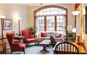 A cozy living room with a large arched window letting in natural light. The room features a floral-patterned sofa, two red armchairs, a wooden coffee table, a small side table with a lamp, and a wooden chair in the foreground. The walls are decorated with framed pictures and there are floor lamps providing additional lighting.