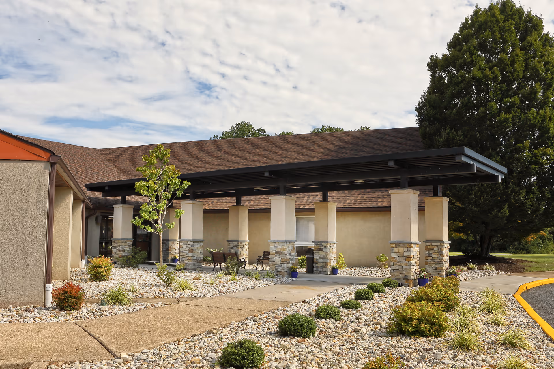 Covered entrance/porte-cochère with stone pillars, walkway, and landscaped rock beds in front of a single-story building.