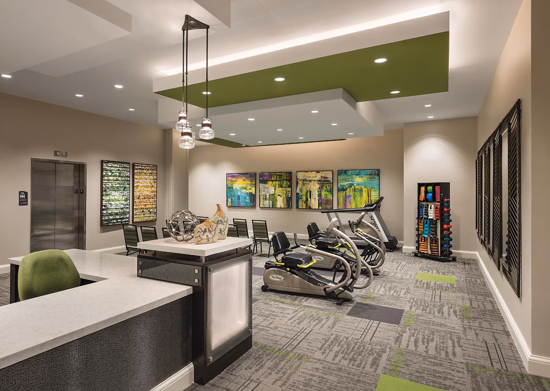A modern fitness room with exercise equipment including stationary bikes and a treadmill. The room features a reception desk with a green chair, decorative sculptures, colorful abstract paintings on the wall, and a rack of various dumbbells. The ceiling has recessed lighting and green accent panels.