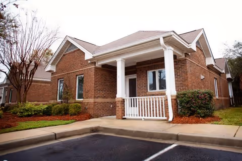 Exterior view of a single-story brick building with a small covered porch supported by white columns. There are shrubs and small trees planted around the building, and a parking area with marked spaces in the foreground.