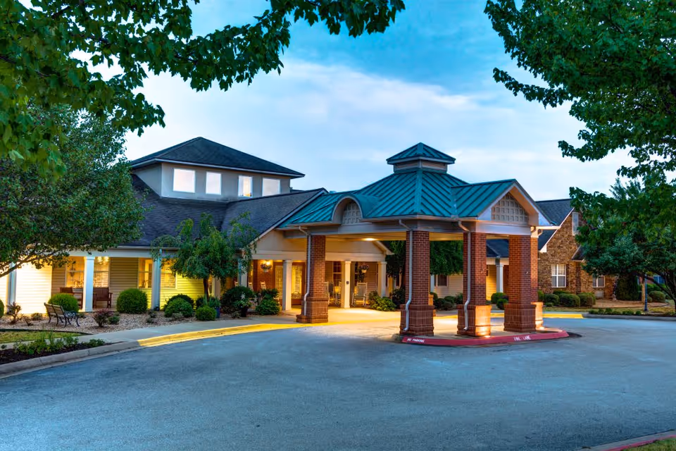 Exterior view of Brookstone Assisted Living Community at dusk, showing a covered entrance with brick pillars and a green metal roof, surrounded by trees and landscaping.