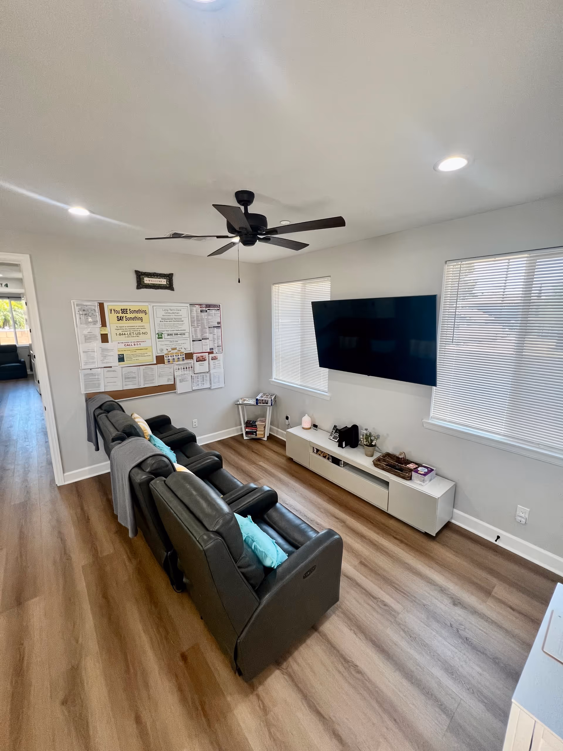 Small common living room with a row of black reclining chairs facing a wall-mounted TV, windows, and a bulletin board.