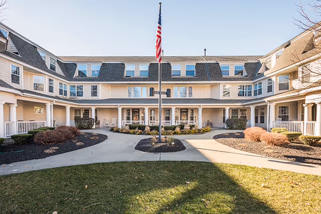 Exterior view of a senior living facility courtyard with a central flagpole flying the American flag. The building is three stories tall with beige siding and white trim, featuring multiple windows and covered walkways with white railings. The courtyard has landscaped areas with shrubs and mulch, and a paved walkway leading to the entrance.