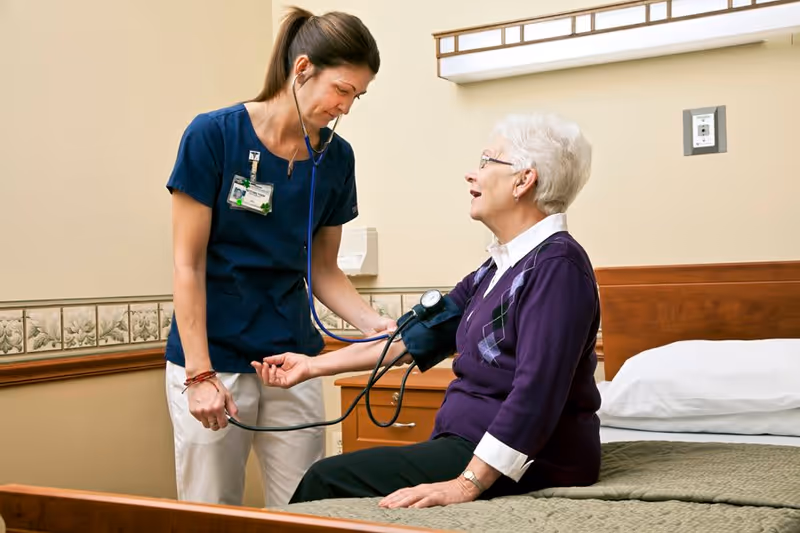 A healthcare professional in navy scrubs is taking the blood pressure of an elderly woman with white hair who is sitting on a bed in a room. The elderly woman is wearing glasses and a purple sweater with an argyle pattern. The room has beige walls and a wooden bed frame with a green bedspread.