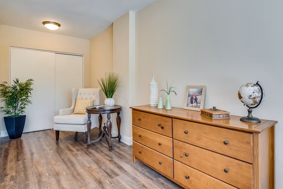 A cozy corner of a senior living facility room featuring a wooden dresser with decorative items including a globe, framed photo, and vases. Next to the dresser is a small round wooden table with a plant and an open book, accompanied by a cushioned armchair with a decorative pillow. A potted plant is placed near a closed white sliding door, and the room has wood flooring and beige walls.