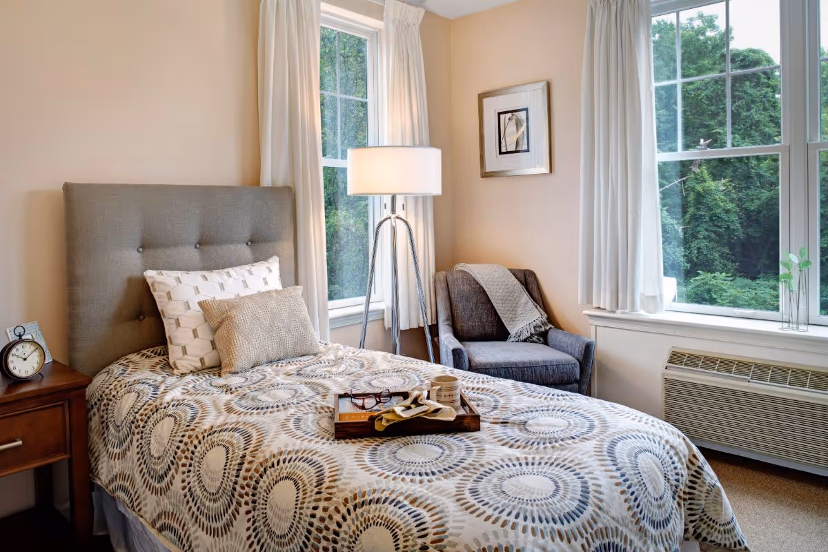 A cozy bedroom with a single bed featuring a patterned bedspread and two pillows. A wooden nightstand with an alarm clock is beside the bed. There is a gray upholstered armchair with a throw blanket next to a floor lamp. Two large windows with white curtains provide a view of green trees outside. A framed picture hangs on the wall above the armchair.