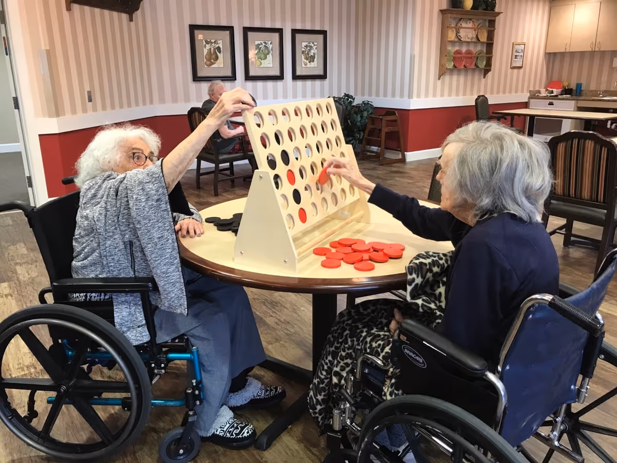 Two elderly women in wheelchairs playing a large wooden Connect Four game at a round table in a common area with striped wallpaper, framed pictures, and wooden flooring.