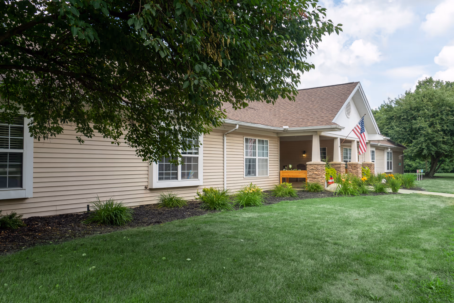 Exterior view of a single-story building with beige siding and a brown shingled roof, surrounded by green grass and trees. The building has multiple windows and a covered porch with stone pillars, an American flag, and some plants near the entrance.