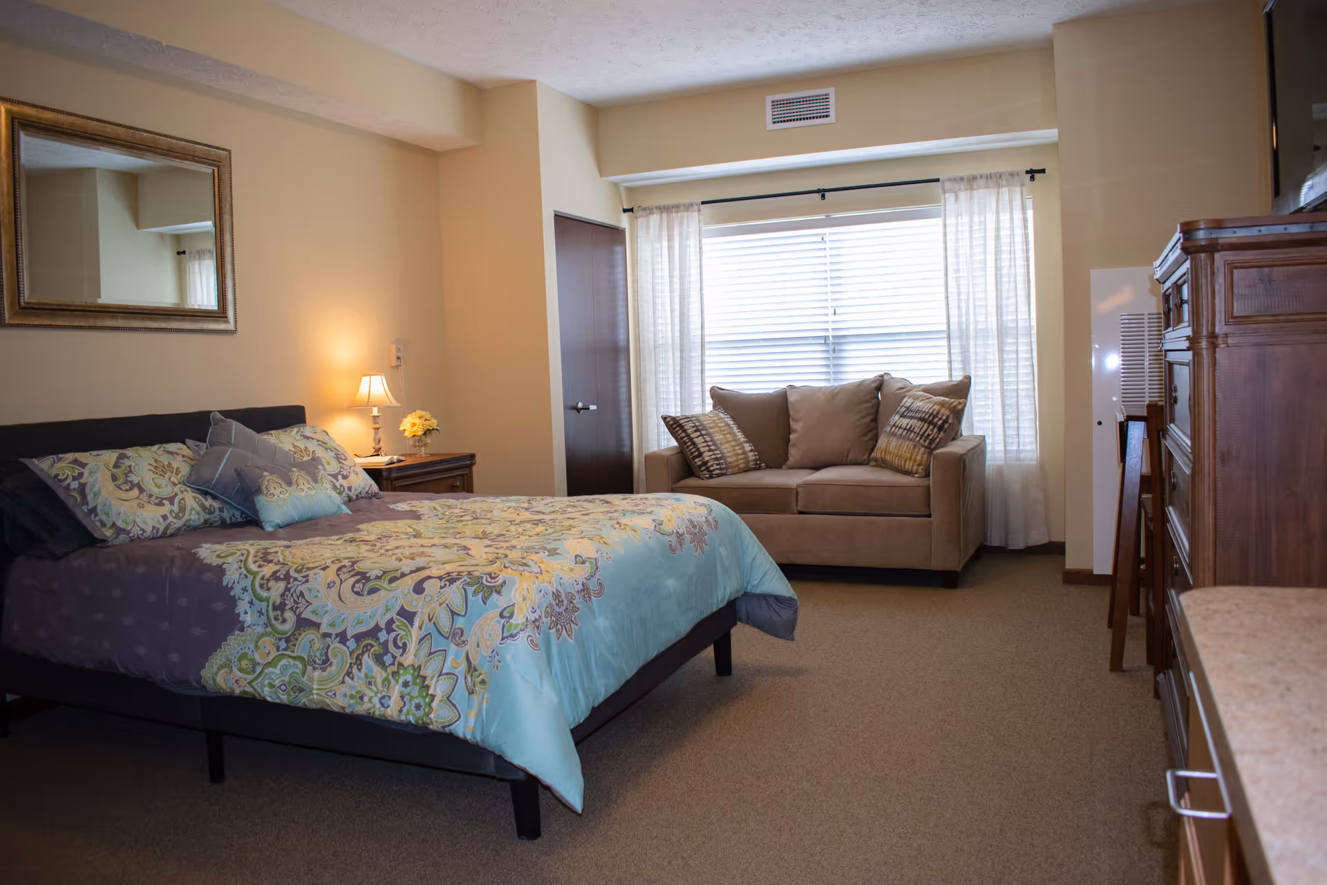 A bedroom with a bed covered in a colorful patterned comforter and pillows, a nightstand with a lamp and flowers, a large mirror on the wall, a beige sofa with patterned cushions near a window with white curtains, and wooden furniture including a dresser and a cabinet.