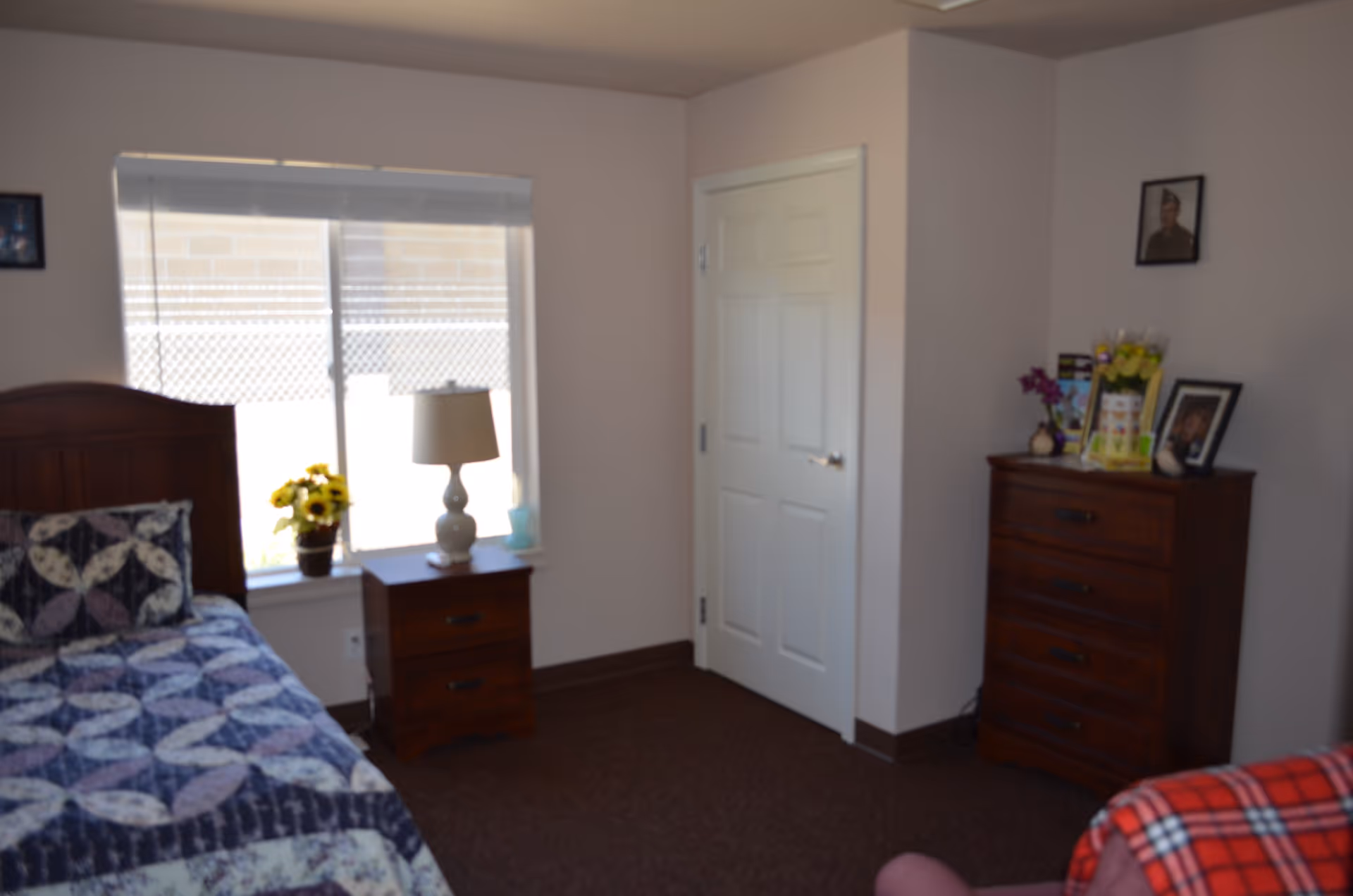 A bright bedroom with a patterned quilted bed, a nightstand with a lamp and flowers by a window, and a wooden dresser against the wall.
