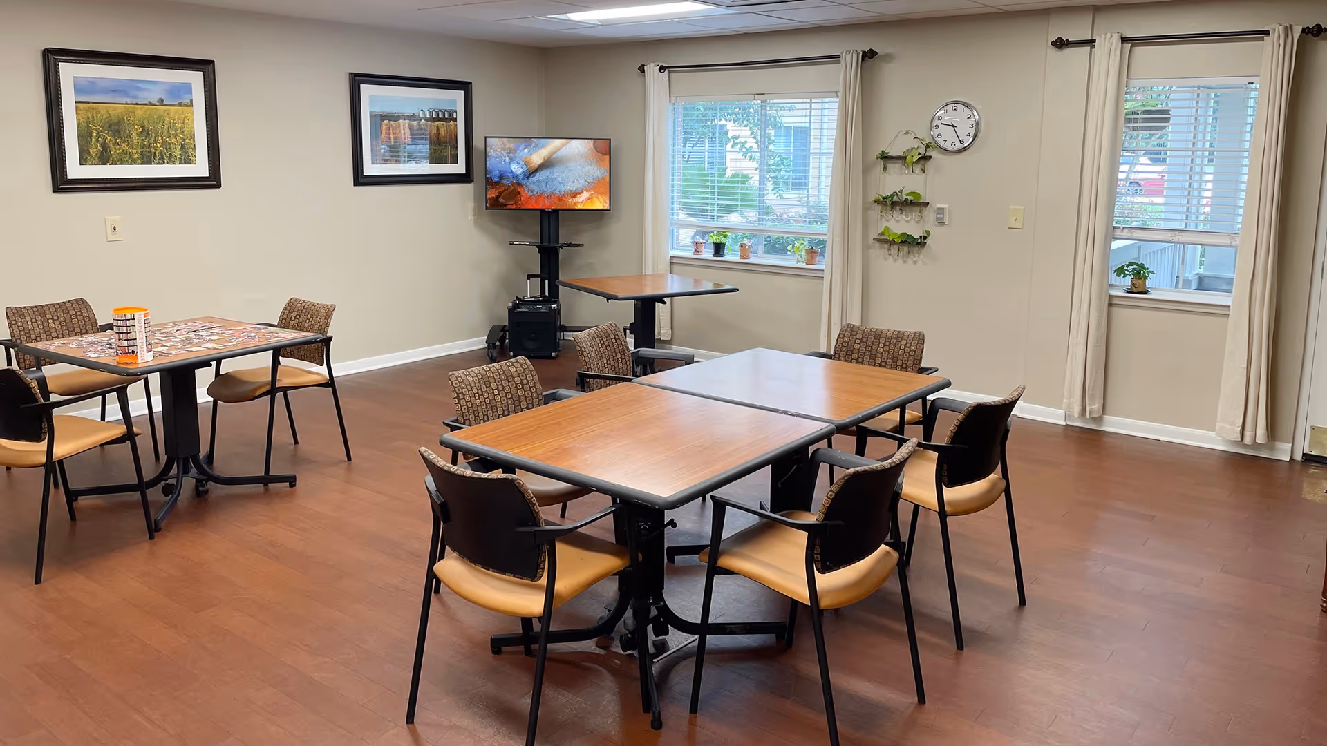 A well-lit room with wooden flooring featuring two tables with chairs around them. One table has a puzzle on it. There are two framed landscape pictures on the wall, a TV on a stand, windows with beige curtains, a wall clock, and small potted plants on the windowsill and wall-mounted shelves.