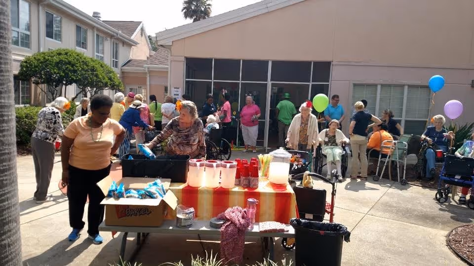 A group of elderly people and caregivers gathered outdoors in a courtyard area of a senior living facility. There is a table in the foreground with pitchers of drinks, cups, and snacks. Some people are sitting, some are standing, and a few are using walkers or wheelchairs. Colorful balloons are tied to some chairs and walkers, creating a festive atmosphere.