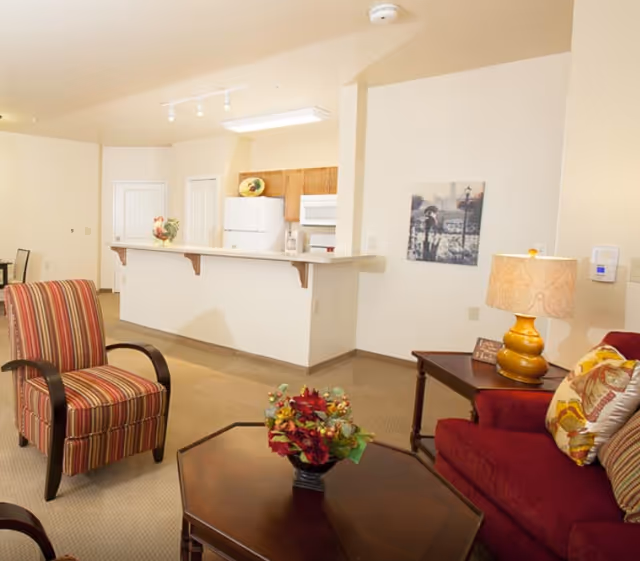Open living area with upholstered chairs, a coffee table with a floral arrangement, and a kitchen with a breakfast bar.