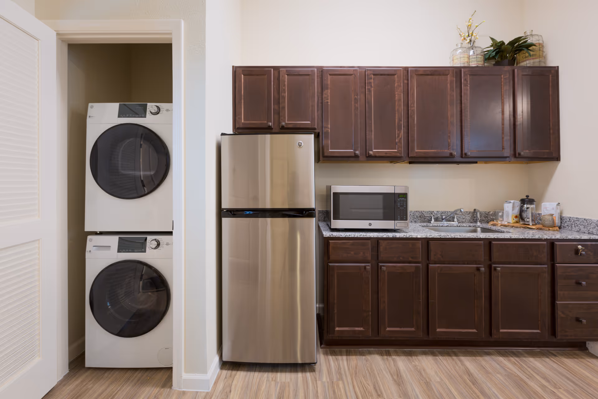 A compact kitchen area with dark wooden cabinets, a stainless steel refrigerator, a microwave on the countertop, and a sink. To the left, there is a small laundry closet with a stacked washer and dryer. The floor has a light wood finish.