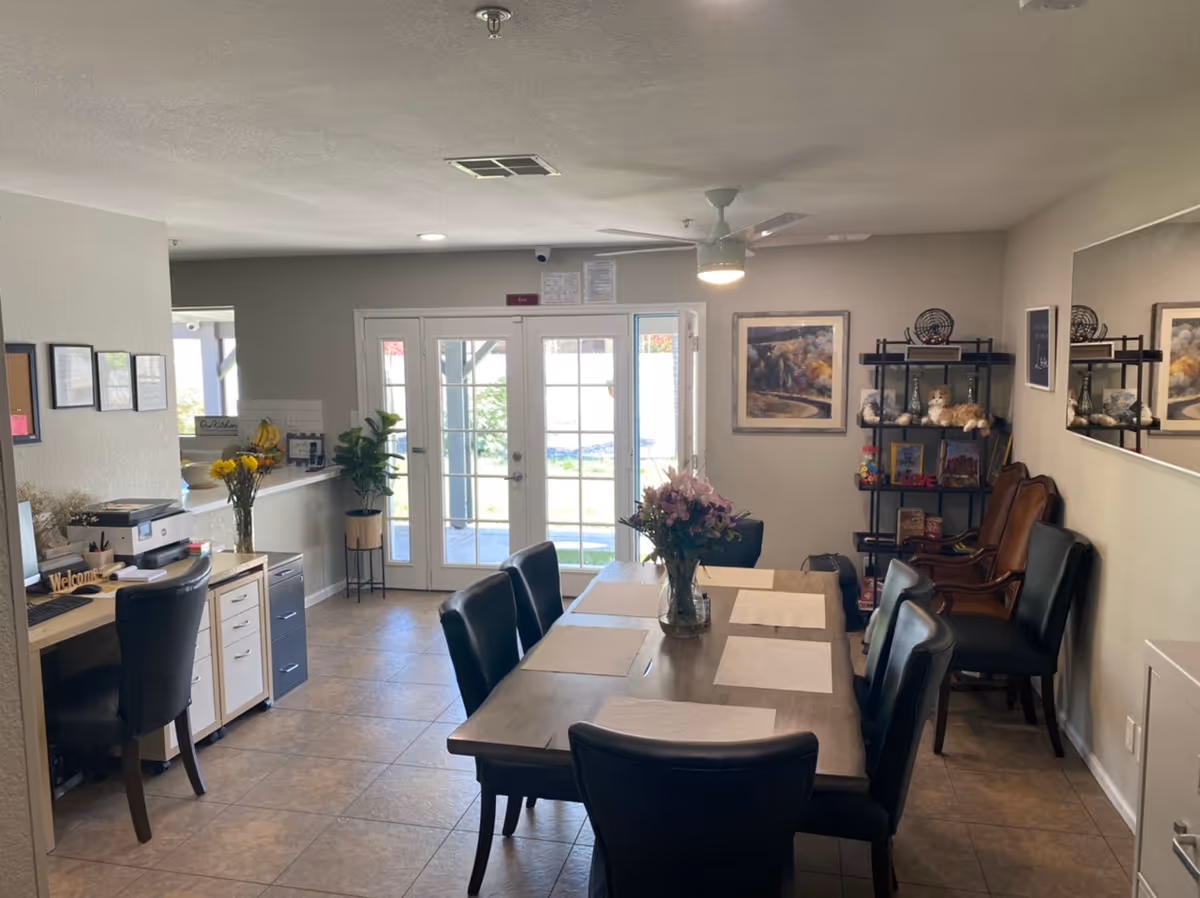 Interior view of a dining room in an assisted living home with a wooden dining table set with six black chairs and placemats. A vase with flowers is placed in the center of the table. In the background, there are glass double doors leading outside, a small office desk with a chair and printer on the left, and a shelving unit with decorative items and books on the right. The room has tiled flooring, neutral-colored walls, and a ceiling fan with a light.