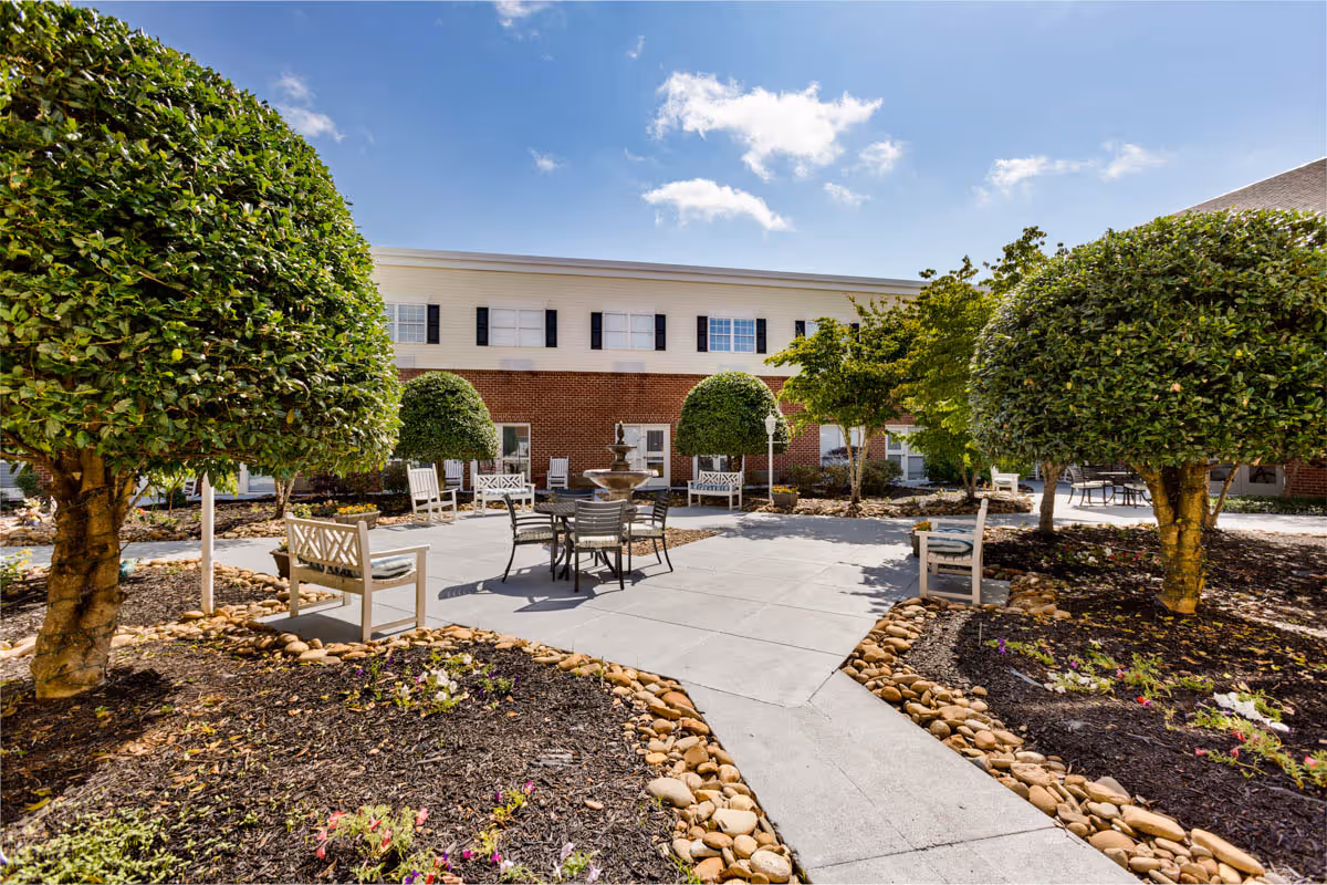 Sunlit courtyard with benches, tables, a central fountain and trimmed topiary trees in front of a two-story brick-and-siding building.