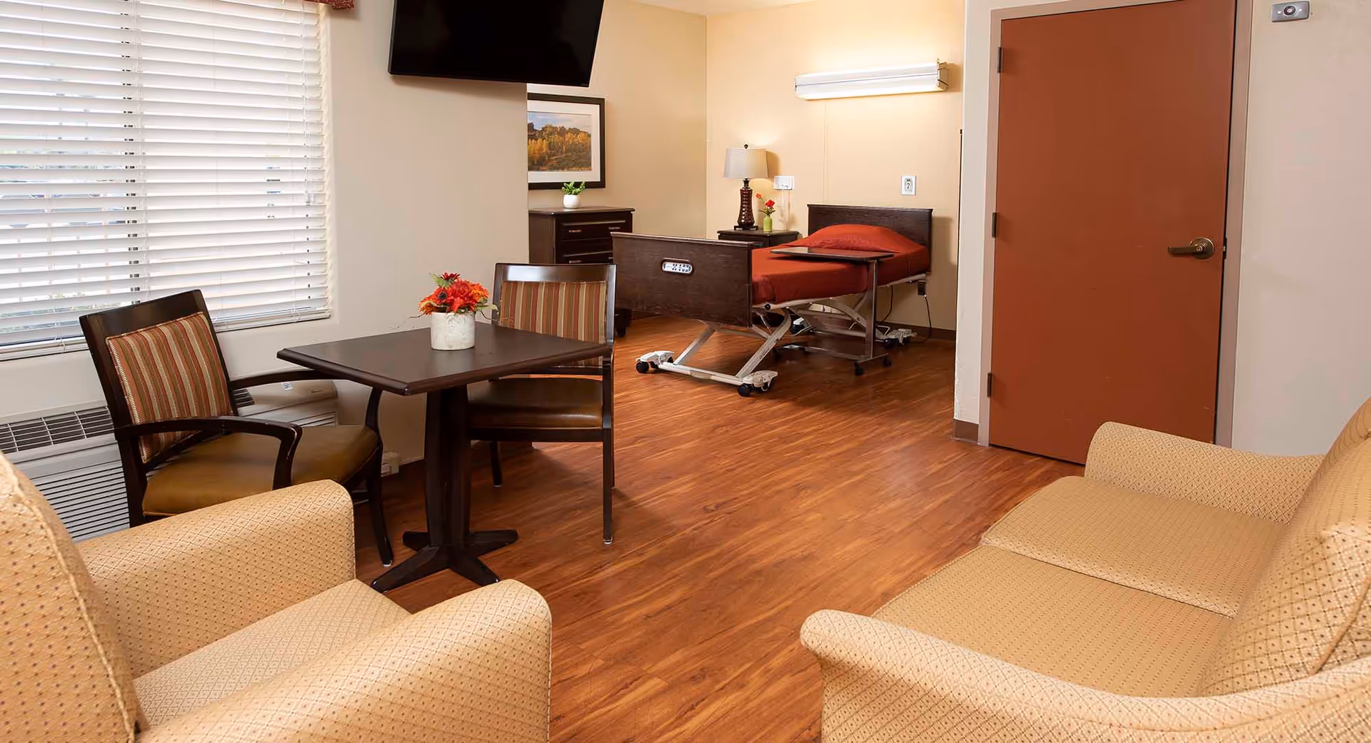 A senior care facility room with a hospital-style bed with a red blanket, a small table with two chairs, a beige armchair and sofa, a dresser, a wall-mounted TV, and a window with blinds. The room has wood flooring and beige walls.