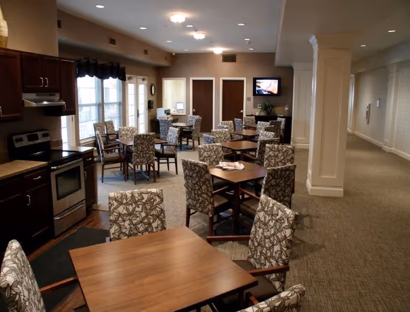 Interior view of a dining area in a senior living facility with multiple wooden tables and cushioned chairs featuring a leaf pattern. There is a kitchen area with stainless steel appliances on the left side, large windows with dark valances, and a hallway extending to the right. A small television is mounted on the far wall above a cabinet.